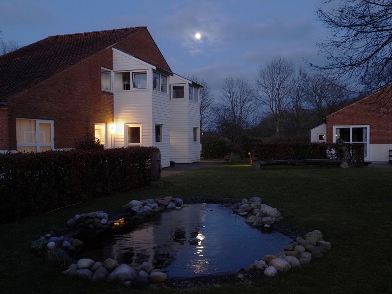 Aftenfoto af fællesplæne med dam og to huse i baggrunden.